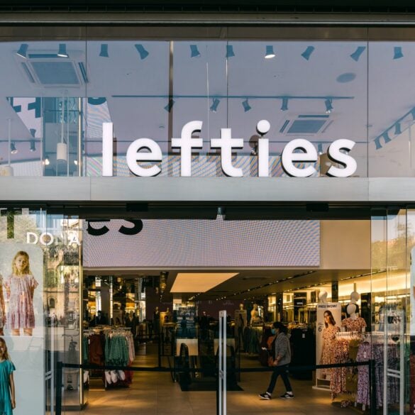 Glasfassade einer Lefties-Filiale in Badalona bei Barcelona mit beleuchtetem Schriftzug über dem Eingang, im Schaufenster hängen Plakate mit Kindermode, dahinter der Verkaufsraum.