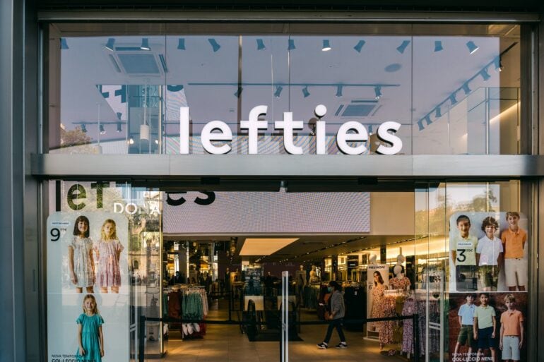 Glasfassade einer Lefties-Filiale in Badalona bei Barcelona mit beleuchtetem Schriftzug über dem Eingang, im Schaufenster hängen Plakate mit Kindermode, dahinter der Verkaufsraum.
