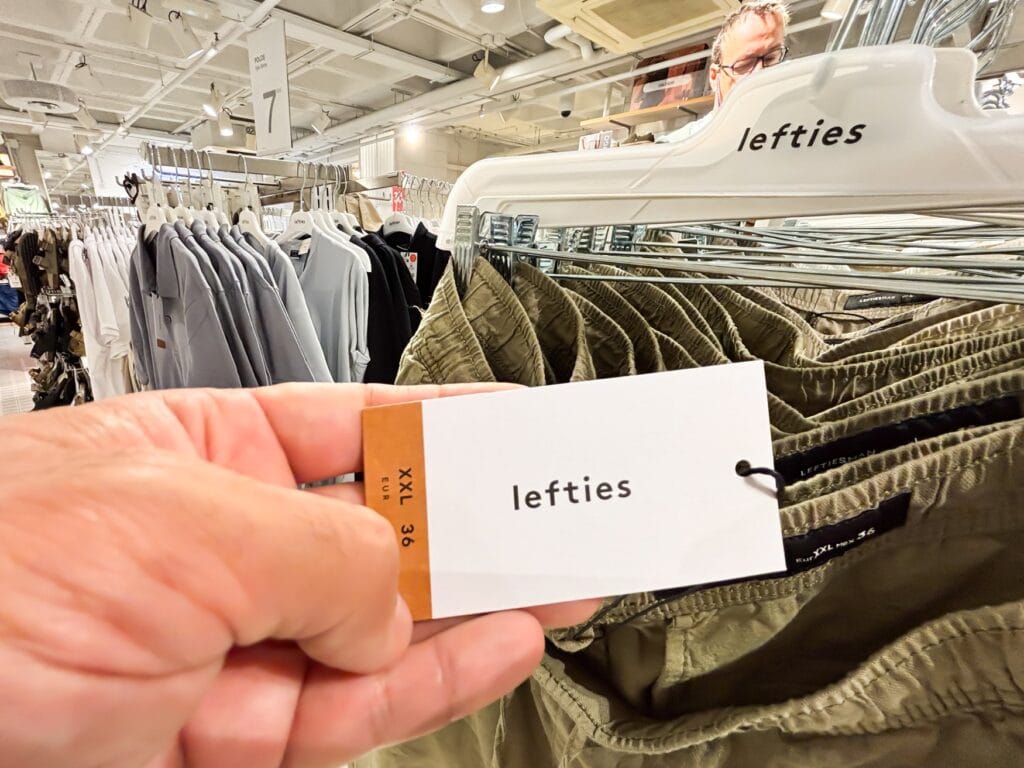 Eine Hand hält ein Preisschild mit Lefties-Logo vor eine Reihe olivgrüner Hosen an einer Kleiderstange, im Hintergrund Regale und weitere Ware im Verkaufsraum.