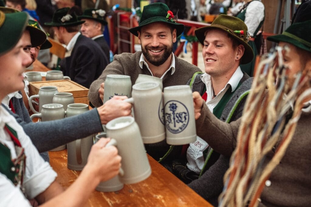 Männer in Trachtenhüten und Janker stoßen an einem Holztisch mit steinernen Bierkrügen an.