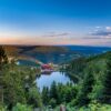 Der Mummelsee im Schwarzwald liegt in einer bewaldeten Mulde, am Ufer steht ein Berghotel mit rotem Dach. Von einem Aussichtspunkt zwischen Tannen reicht der Blick über die Höhenzüge bis zum Horizont.