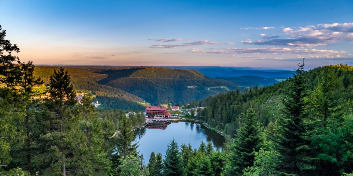 Der Mummelsee im Schwarzwald liegt in einer bewaldeten Mulde, am Ufer steht ein Berghotel mit rotem Dach. Von einem Aussichtspunkt zwischen Tannen reicht der Blick über die Höhenzüge bis zum Horizont.