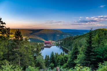 Der Mummelsee im Schwarzwald liegt in einer bewaldeten Mulde, am Ufer steht ein Berghotel mit rotem Dach. Von einem Aussichtspunkt zwischen Tannen reicht der Blick über die Höhenzüge bis zum Horizont.