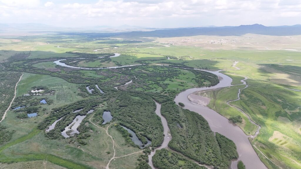 Luftaufnahme des Orchontals in der Mongolei. Der Fluss mäandert in weiten Schleifen durch grüne Auwälder und Altarme, dahinter öffnet sich die Steppe mit einer Ortschaft und Hügelketten.