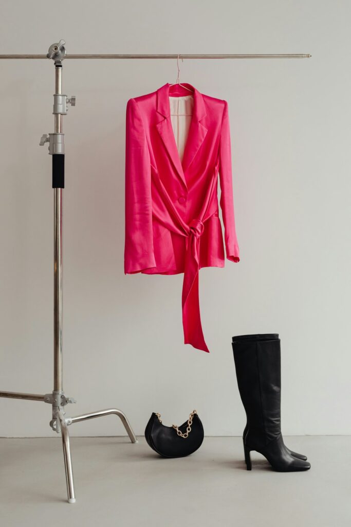 Ein pinkfarbener Satin-Blazer mit Bindegürtel hängt an einem Studiostativ, davor stehen schwarze Lederstiefel und eine schwarze Handtasche mit Goldkette.