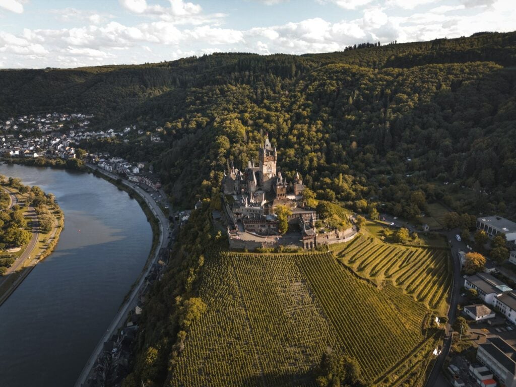 Luftaufnahme der Reichsburg Cochem auf einem bewaldeten Felssporn über der Mosel. Unterhalb der Burg ziehen sich Weinbergszeilen den Hang hinab, links folgt der Fluss einer weiten Kurve.