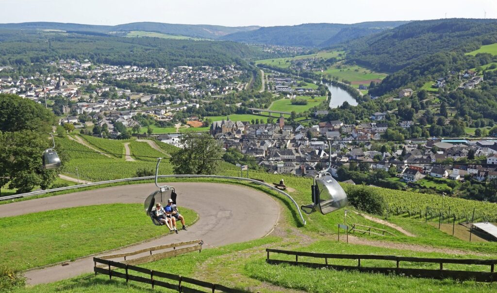 Ein Paar fährt in einem offenen Sessel der Saarburger Sesselbahn über eine Wiese talwärts, ein leerer Sessel kommt entgegen. Unten liegen die Stadt Saarburg, die Saar und Weinberge.