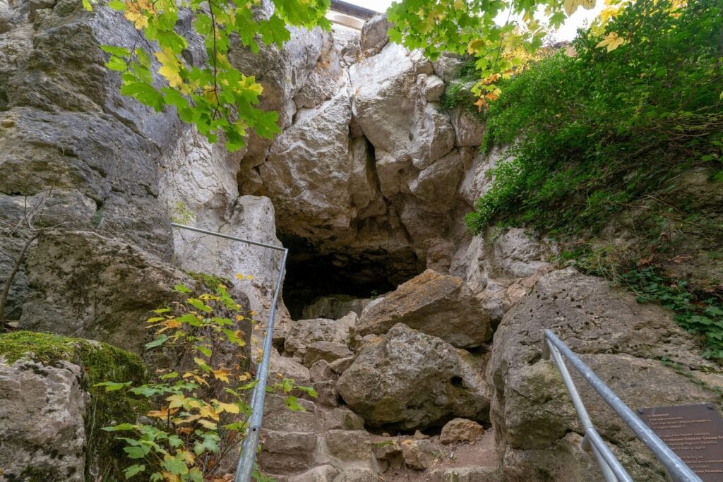 Der Eingang zur Höhle Sibyllenloch unterhalb der Burg Teck bei Owen. Zwischen hellen Kalkfelsen und herbstlich gefärbtem Laub führen Stufen mit Metallgeländer in den dunklen Spalt.