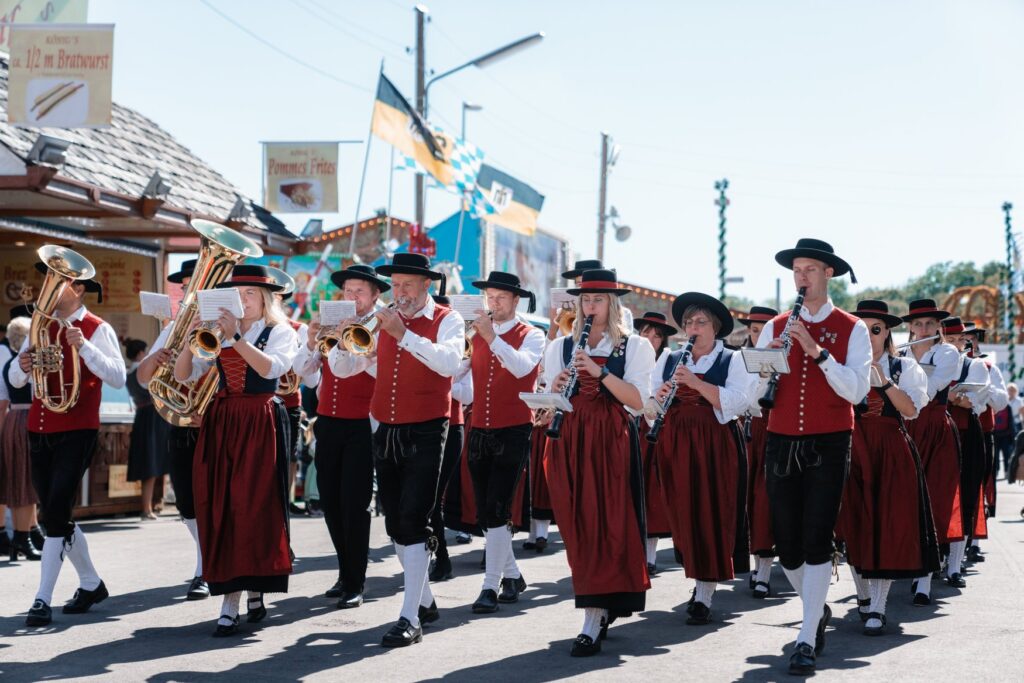 A band in red waistcoats and dirndls plays during the parade across the Theresienwiese, with flags and food stalls behind.