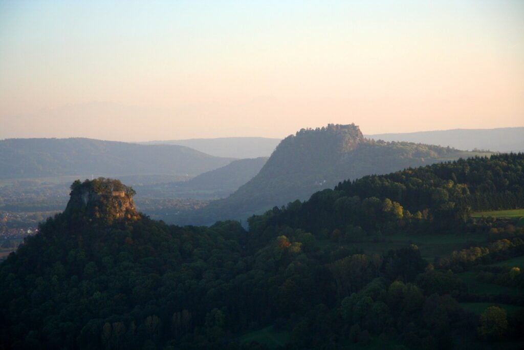 Blick über die Vulkanlandschaft des Hegau im Abendlicht. Im Vordergrund erhebt sich ein bewaldeter Kegelberg mit Mauerresten auf dem Felsplateau, dahinter liegt der Hohentwiel mit seiner Festungsruine im Dunst.