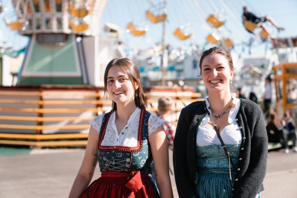 Zwei lachende Frauen im Dirndl gehen über die Wiesn, hinter ihnen dreht sich ein Kettenkarussell.