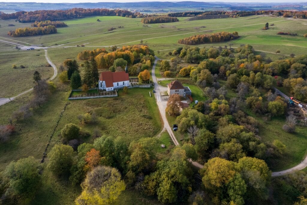 Lost Places in Baden-Württemberg: Luftaufnahme der Kirche und des ehemaligen Schulhauses von Gruorn, umgeben von Wiesen, Wegen und herbstlich gefärbten Bäumen.