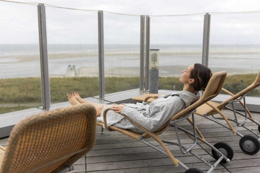 Eine Frau im grauen Bademantel liegt auf einer Rattanliege auf der Terrasse des Gezeitenlands und hat die Augen geschlossen. Hinter der Glasbrüstung reicht der Blick über die Düne auf Strand und Meer.
