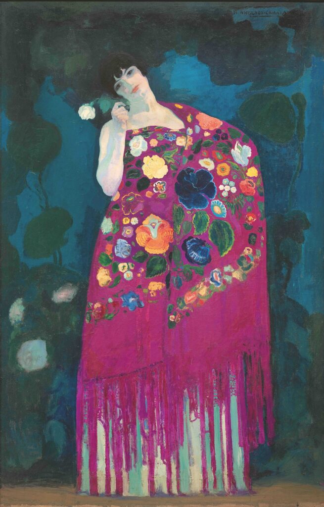 Hermen Anglada Camarasa, „Frau aus Granada”, um 1914: Eine Frau in einem pinkfarbenen Manila-Schal mit aufgestickten Blüten und langen Fransen steht vor dunkelblaugrünem Grund und hält eine weiße Rose ans Gesicht.