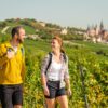 Wandern in Rheinhessen: Zwei Wandernde mit Rucksäcken auf einem Weg durch die Weinberge bei Oppenheim. Im Hintergrund ist die Katharinenkirche zu sehen.