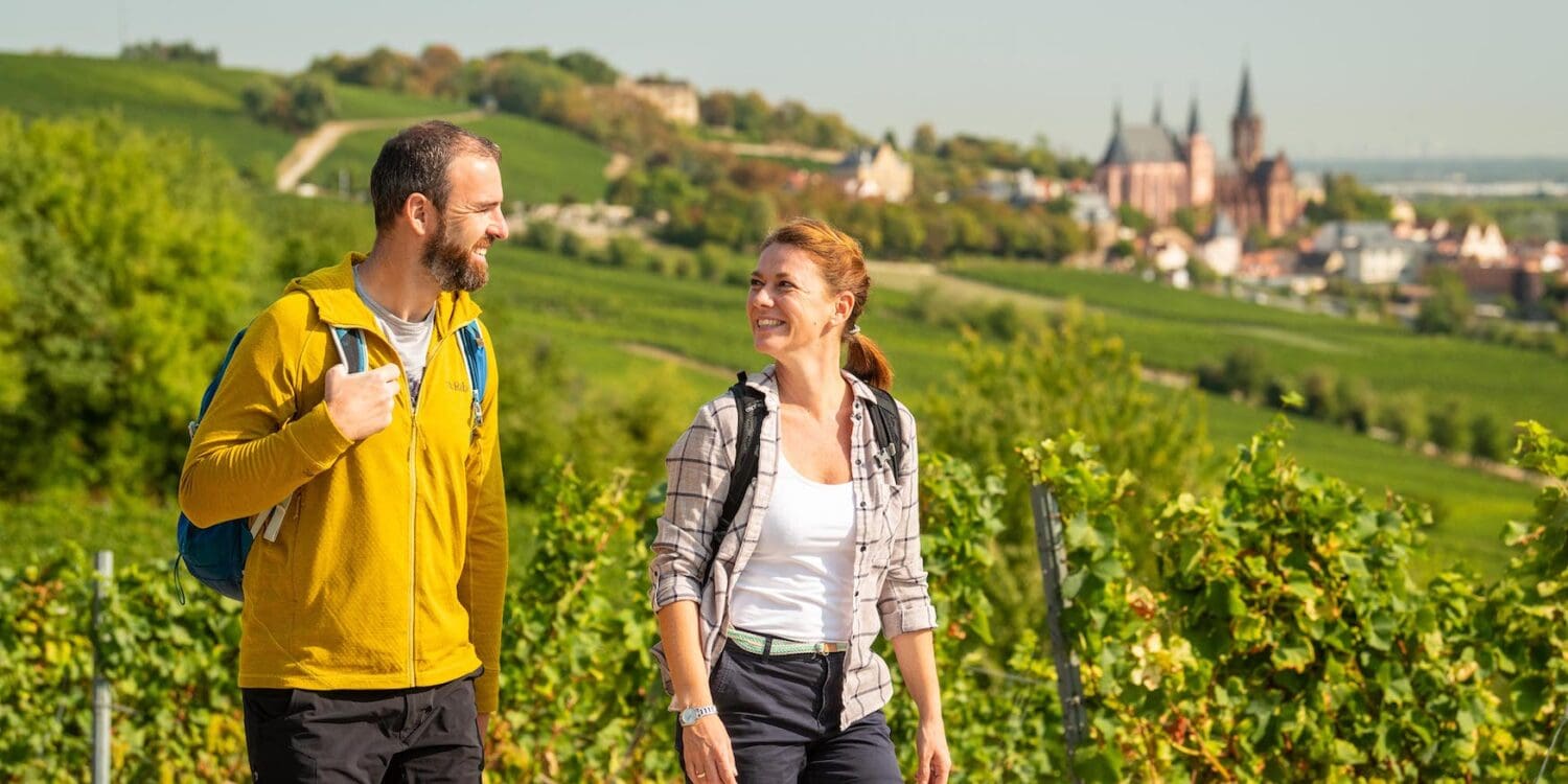 Wandern in Rheinhessen: Zwei Wandernde mit Rucksäcken auf einem Weg durch die Weinberge bei Oppenheim. Im Hintergrund ist die Katharinenkirche zu sehen.
