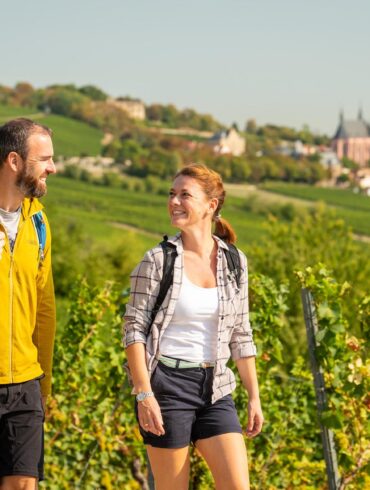Wandern in Rheinhessen: Zwei Wandernde mit Rucksäcken auf einem Weg durch die Weinberge bei Oppenheim. Im Hintergrund ist die Katharinenkirche zu sehen.