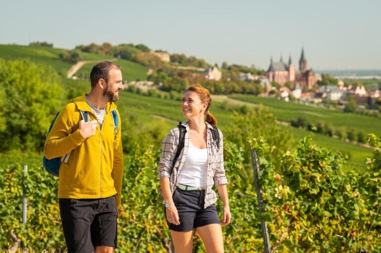 Wandern in Rheinhessen: Zwei Wandernde mit Rucksäcken auf einem Weg durch die Weinberge bei Oppenheim. Im Hintergrund ist die Katharinenkirche zu sehen.