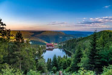 View over a dark forested valley in the Black Forest to a small lake. A hotel with a red roof stands at the shore and is mirrored in the still water, the sky above turning pink at sunset.