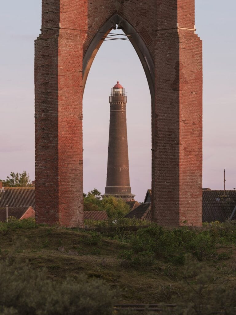 Blick durch den Spitzbogen eines Backsteinbauwerks auf den Neuen Leuchtturm von Borkum. Der dunkle Turm mit roter Kuppel steht hinter den Ziegeldächern des Ortes im Abendlicht.